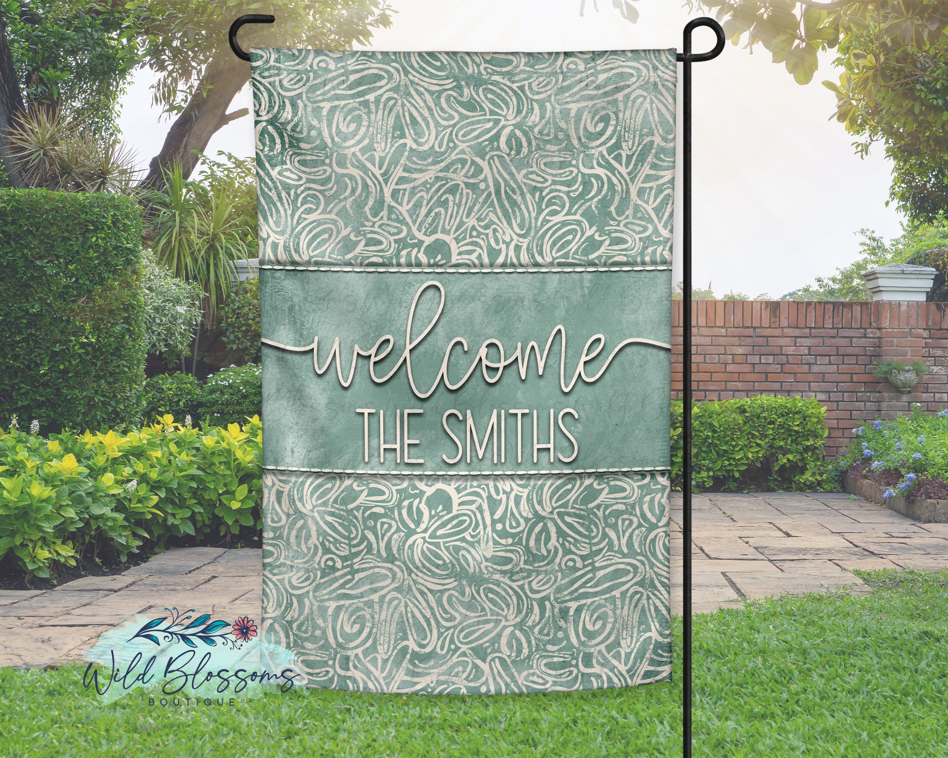 Decorative garden flag with 'Welcome The Smiths' text and floral patterns in a garden setting.