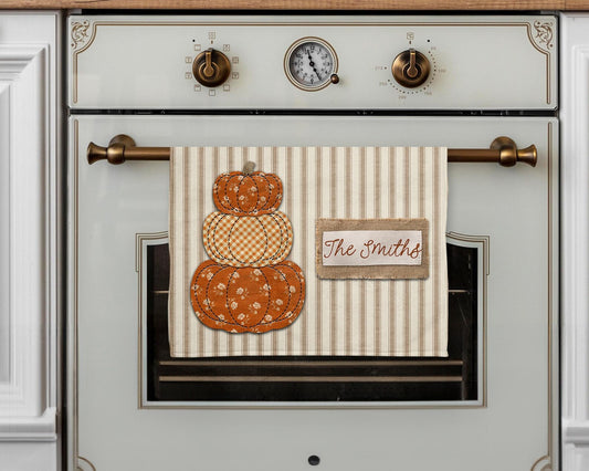 Personalized fall kitchen towel featuring three stacked orange patchwork pumpkins with floral and gingham patterns, stitched details, beige ticking stripe background, and a burlap-style family name label.