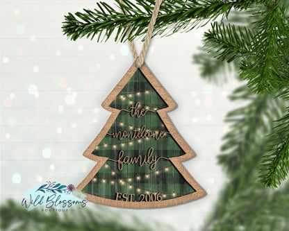 A buffalo plaid Christmas tree ornament with a family name written on it, hanging against a green Christmas tree background.