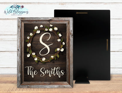 A wooden frame with a black back, featuring a white cotton wreath and the personalized family name 'The Smiths' printed in the center.