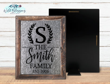 A personalized family name sign with 'The Smith Family' text, featuring a black frame and a gray metallic finish on a wooden background. The sign is rectangular with rounded corners and a visible hinge on the back for easy hanging.